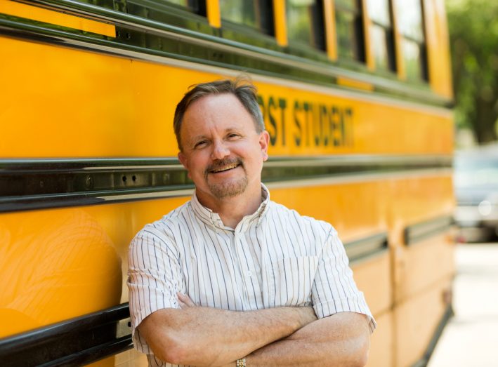 Portrait of a school bus driver standing next to a school bus smiling at camera.